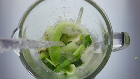 Clear-water-pouring-blender-on-green-raw-vegetables-fruits-close-up-top-view.