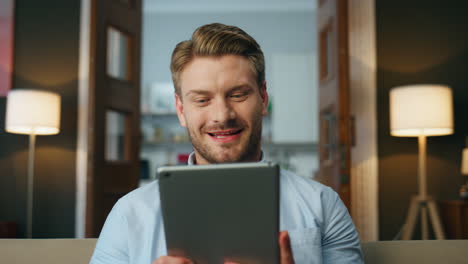 Smiling-guy-watching-tablet-screen-evening-room.-Closeup-excited-man-achieving