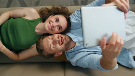 Funny-couple-taking-selfie-laying-couch-top-view.-Happy-man-photographing-tablet