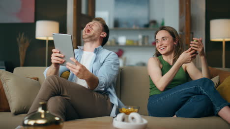 Happy-woman-talking-husband-pointing-tablet-indoors.-Handsome-man-laughing-joke
