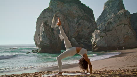 Girl-training-upward-bow-pose-with-leg-raised-up-on-beach.-Woman-practicing-yoga