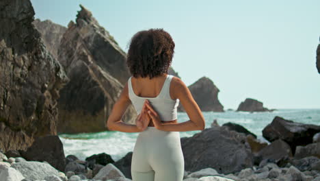 Girl-making-Namaste-hands-behind-back-on-rocky-seashore.-Woman-stretching-arms.