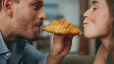 Romantic-pair-eating-croissant-home-zoom-on.-Loving-man-woman-face-bitting-bun