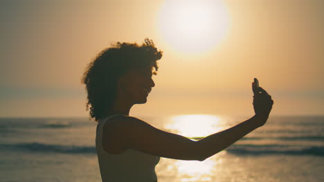 Girl-smiling-phone-camera-standing-beach-at-sunrise-closeup.-Woman-making-selfie