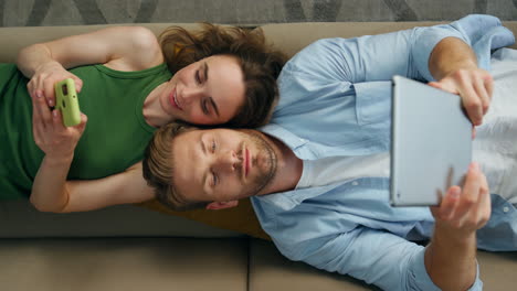 Closeup-chilling-duet-laying-sofa-home.-Involved-couple-using-gadgets-top-view
