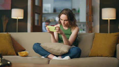 Involved-gamer-playing-cellphone-sofa-room.-Carefree-woman-enjoying-video-games