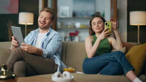 Positive-couple-using-gadgets-at-home-sofa.-Laughing-man-showing-tablet-to-wife