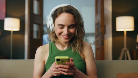 Relaxed-lady-listening-headset-music-at-sofa.-Smiling-girl-turning-smartphone