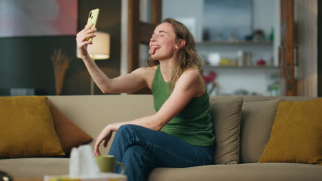 Carefree-woman-showing-tongue-to-smartphone-camera-home.-Happy-lady-funny-face