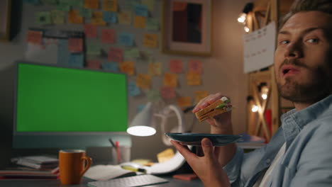 Hungry-student-eating-sandwich-evening-room.-Man-taking-break-in-front-computer