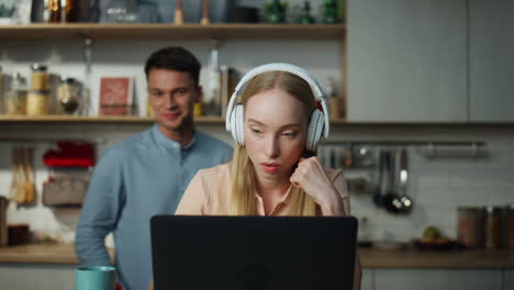 Woman-watching-internet-video-laptop-by-headphones-at-kitchen-with-man-behind.