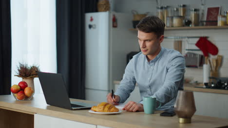 Man-making-notes-working-laptop-in-kitchen.-Freelancer-using-wireless-technology