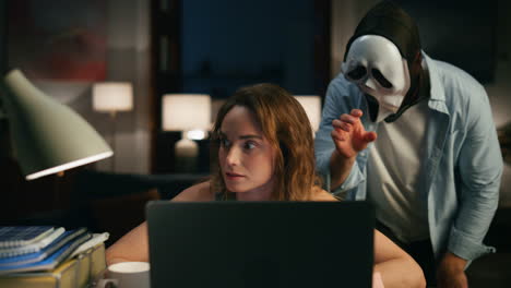 Focused-woman-speaking-laptop-night-closeup.-Masked-man-scream-costume-sneaking