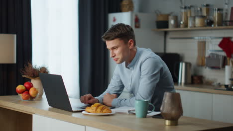 Freelancer-man-satisfied-work-sitting-at-kitchen-table-with-modern-laptop.