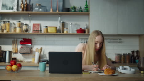 Girl-student-studying-online-sitting-at-kitchen-with-laptop.-Woman-making-notes.