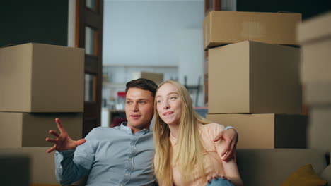 New-owners-sit-down-on-couch-between-carton-boxes-close-up.-Couple-enjoy-moving.