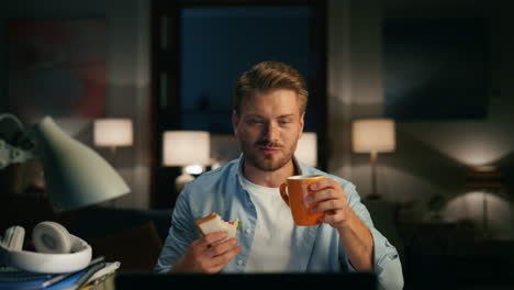 Positive-man-videocalling-home-closeup.-Guy-drinking-taking-sandwich-break