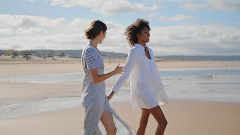 Joyful-lesbian-couple-walking-sea-shore-vertical-video.-Happy-girls-hold-hands