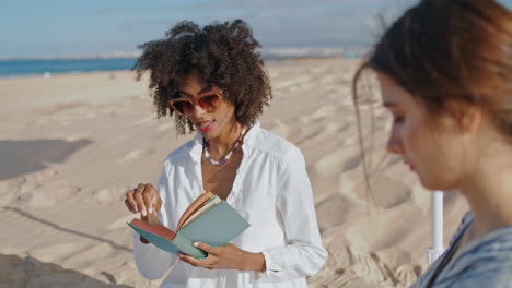 Focused-girl-reading-book-at-ocean-picnic.-Two-friends-resting-on-sandy-beach