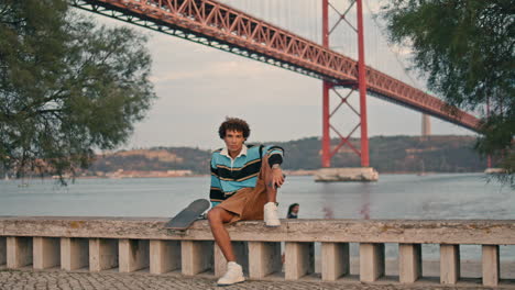 Young-man-sitting-skateboard-at-water-view-portrait.-Skater-guy-looking-camera