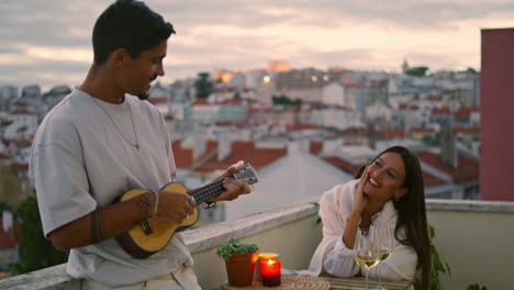 Gutaussehender-Mann,-Der-Auf-Der-Terrasse-Mit-Blick-Auf-Die-Stadt-Bei-Sonnenuntergang-Gitarre-Spielt.-Romantisches-Date-Für-Paare