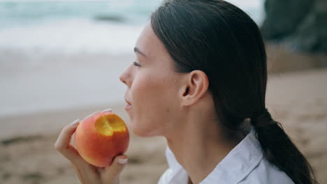 Dreamy-woman-tasting-peach-sitting-on-sand-beach-at-vacation-close-up-vertical