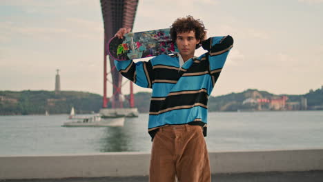 Junger-Mann-Posiert-Mit-Skateboard-Am-Kai-Mit-Blick-Aufs-Wasser.-Skater-Schaut-Vertikal-In-Die-Kamera