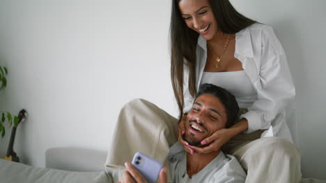 Laughing-woman-kissing-husband-at-couch-closeup.-Married-couple-watching-mobile