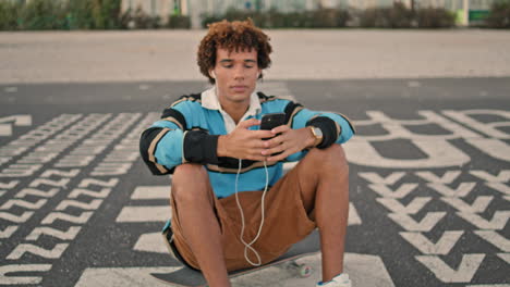 Smiling-guy-enjoying-music-on-phone-outdoors-closeup.-Hipster-sitting-skateboard