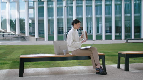 Woman-listening-online-webinar-on-laptop-sitting-bench-near-modern-office.