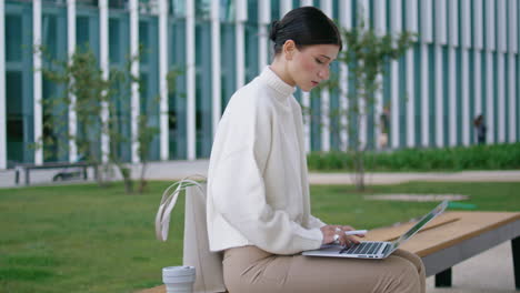Woman-typing-laptop-sitting-on-bench-near-modern-building.-Lady-working-remotely