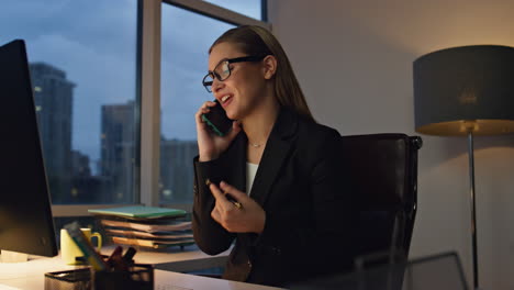 Financial-manager-talking-cellphone-staying-late-in-office.-Smiling-woman-work