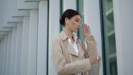 Woman-speaking-wireless-earbuds-standing-near-modern-city-building-close-up.