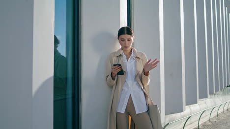 Business-woman-talking-video-conference-by-smartphone-leaning-building-vertical
