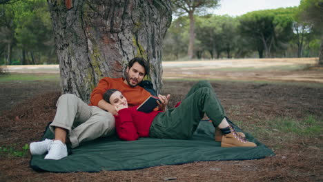 Relaxed-couple-lying-picnic-blanket-tree-vertically.-Man-reading-book-to-woman