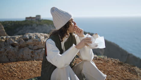 Girl-hiker-eating-food-at-ocean-cliff-vertical.-Serene-travel-breakfast-at-ocean