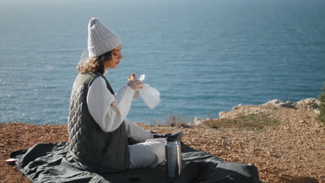 Girl-eating-ocean-picnic-on-cliff-edge-alone.-Serene-tourist-admire-landscape