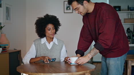 Barista-bringing-cup-coffee-to-african-american-woman-client.-Girl-sitting-table