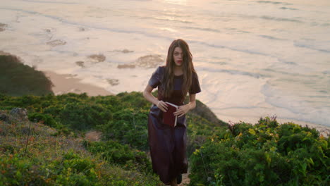 Woman-holding-book-walking-green-hill-in-front-evening-ocean.-Girl-rest-seashore