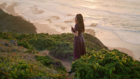 Calm-brunette-relaxing-seashore-standing-hill-holding-book.-Girl-posing-nature.