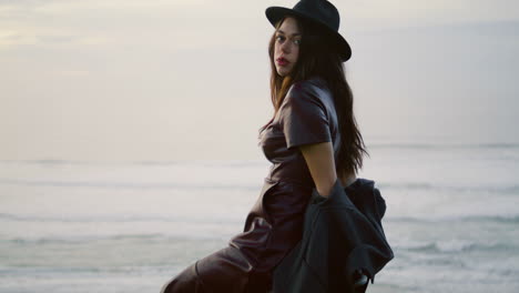 Woman-posing-night-beach-in-elegant-hat.-Girl-sitting-chair-at-gloomy-seascape.