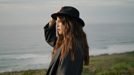 Model-posing-holding-hat-at-ocean-coast-gloomy-evening-close-up.-Serious-woman