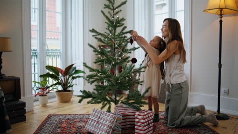 Cheerful-mother-daughter-decorating-Christmas-tree-home.-Mom-child-putting-toys