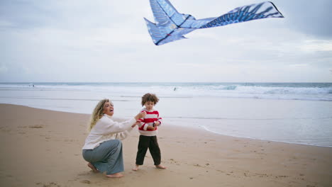 Cheerful-kid-playing-kite-with-mother-on-beach.-Caring-young-parent-helping-son