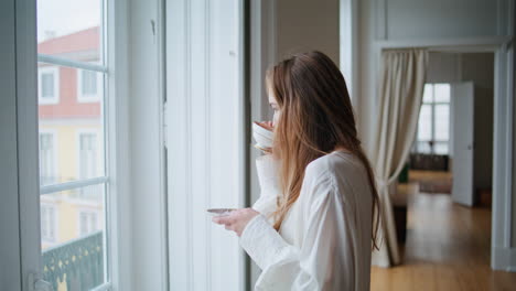 Calm-woman-sipping-coffee-at-flat-interior-closeup.-Girl-watching-window-posing