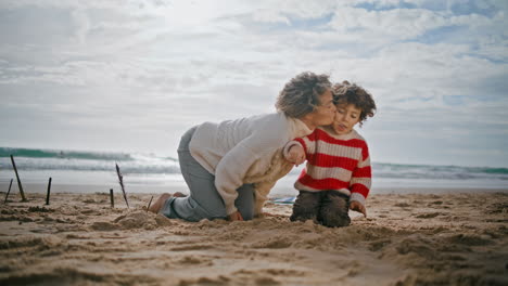 Young-mom-kissing-child-on-weekend-holidays.-Happy-parent-building-sand-castle