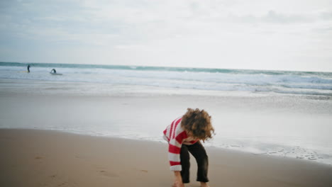 Kid-playing-autumn-beach-with-parent-on-cloudy-day.-Barefoot-curly-boy-resting