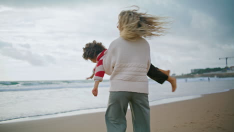 Joyful-mom-spinning-kid-on-autumn-beach.-Smiling-babysitter-playing-airplane