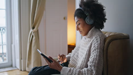Focused-girl-scrolling-pad-comfy-home.-African-headphones-lady-listening-music