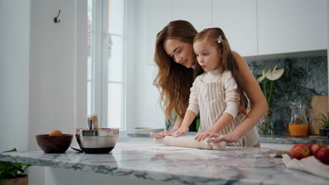 Smiling-mother-teaching-daughter-baking-at-kitchen-interior.-Child-rolling-dough